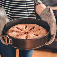 Person holding a baked apple pie in a metal pie dish with oven mitts.