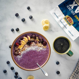 Purple smoothie bowl with toppings on a marble surface with a coffee cup and bag of premium gold natural golden flax.