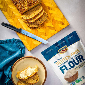 Loaf of bread on a cutting board with a knife, bowl of bread slices, and a bag of Premium Gold gluten free flour on a gray surface.