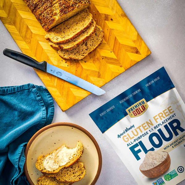 Loaf of bread on a cutting board with a knife, bowl of bread slices, and a bag of Premium Gold gluten free flour on a gray surface.