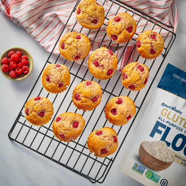 Muffins with raspberries on a cooling rack next to a bag of premium gold brand gluten free all purpose flour